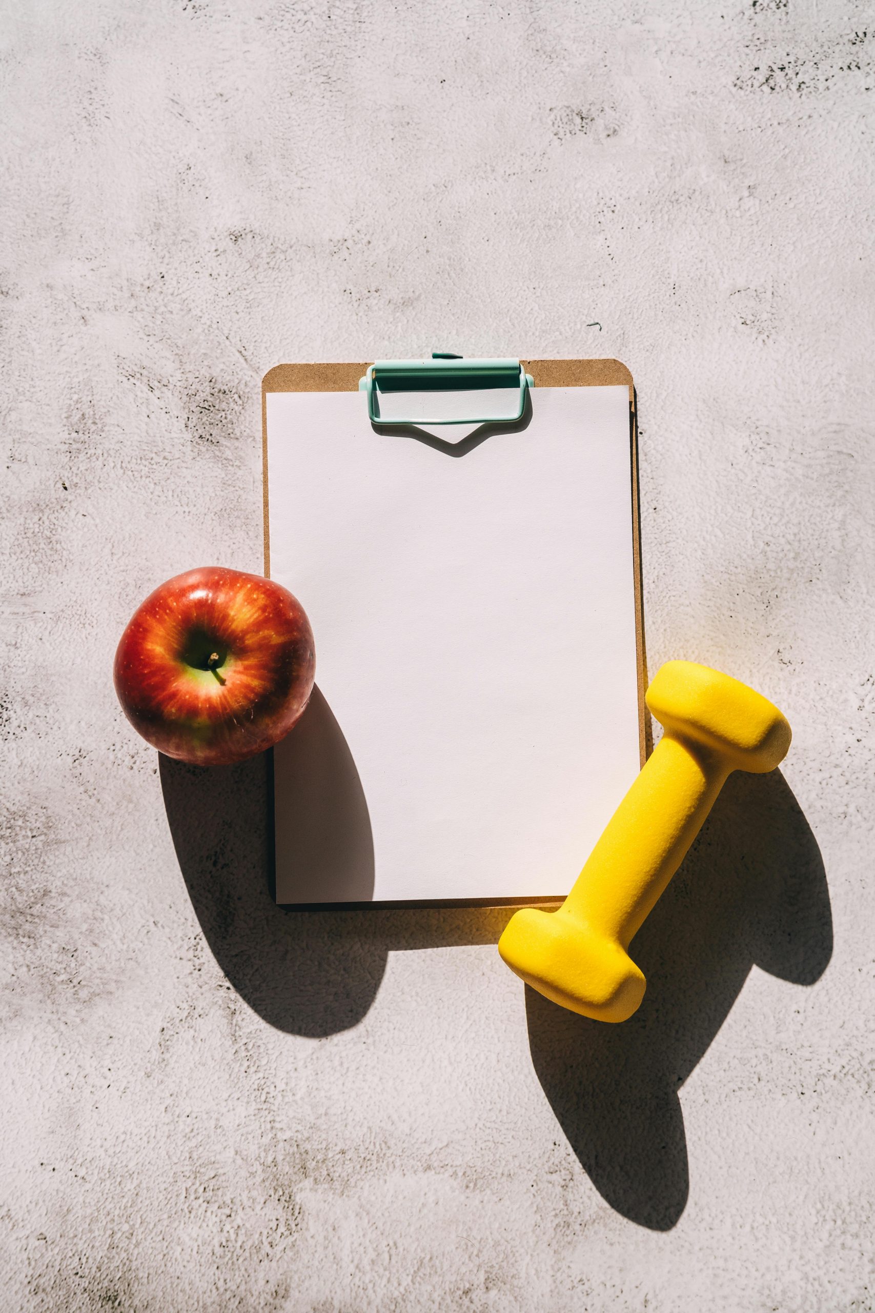 Flat lay of apple, clipboard, and dumbbell representing fitness and nutrition concept.